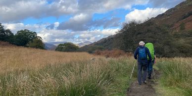 Two hikers walking on a trail surrounded by grass and hills under a partly cloudy sky.