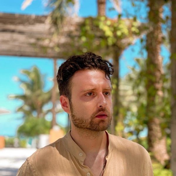 Man with short curly hair and beard in beige shirt outdoors.