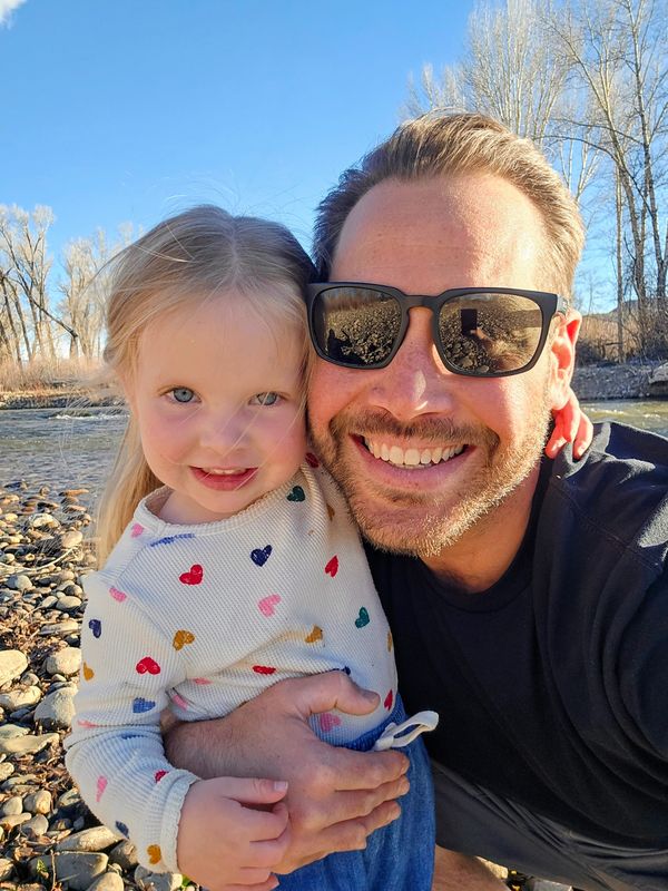 A smiling man with sunglasses hugs a little girl in a heart-patterned sweater by a riverbank.