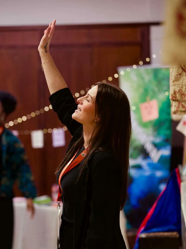 Woman raising her hand, smiling in a lively indoor setting.