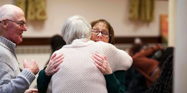 Two elderly women warmly hugging each other indoors.