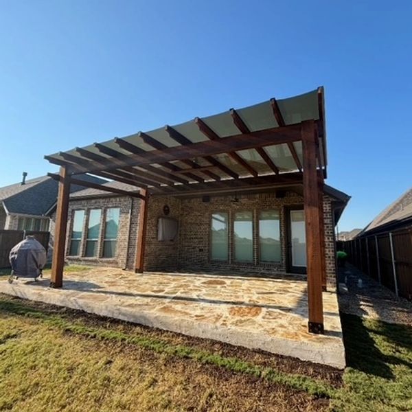Stone patio with wooden pergola attached to a brick house under clear blue sky.