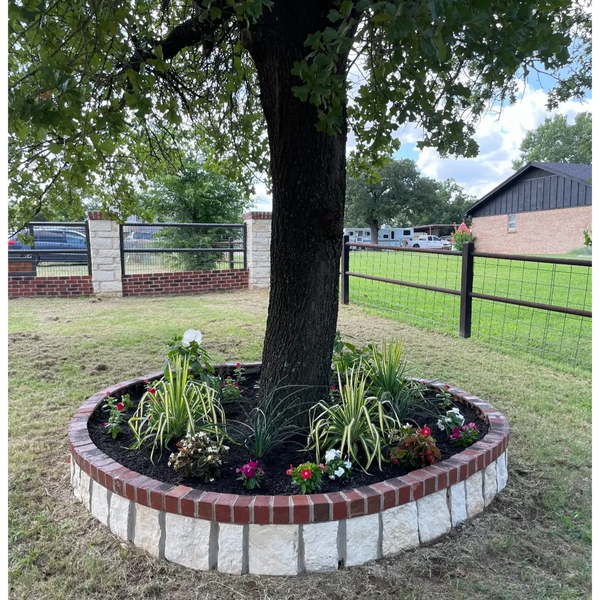 Circular brick and stone planter around a tree with various plants and flowers.