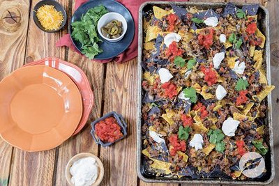 A tray of loaded nachos with toppings and empty plates on a wooden table.