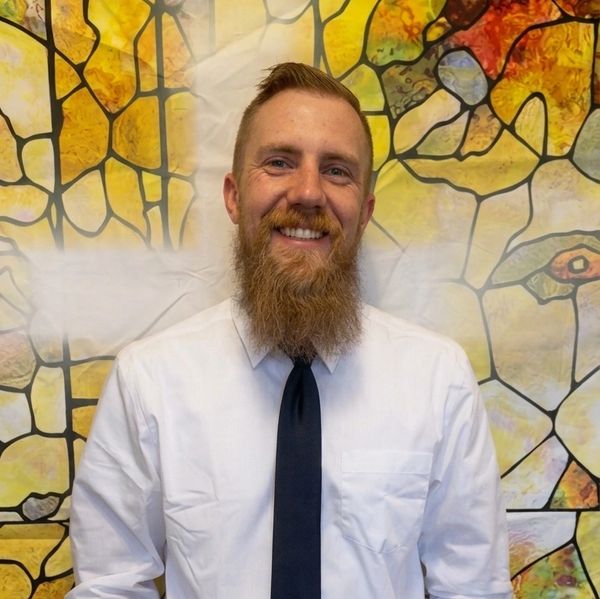 Smiling man with a beard in a white shirt and black tie against a colorful stained glass backdrop.