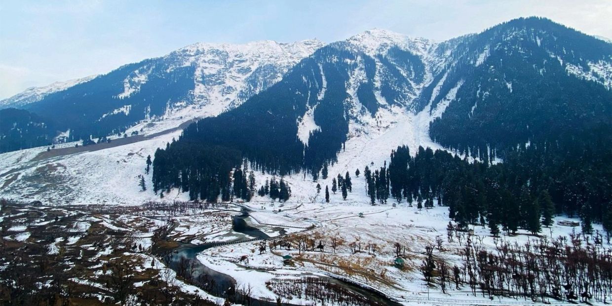 Snow-covered mountains with winding river and scattered trees under clear sky.