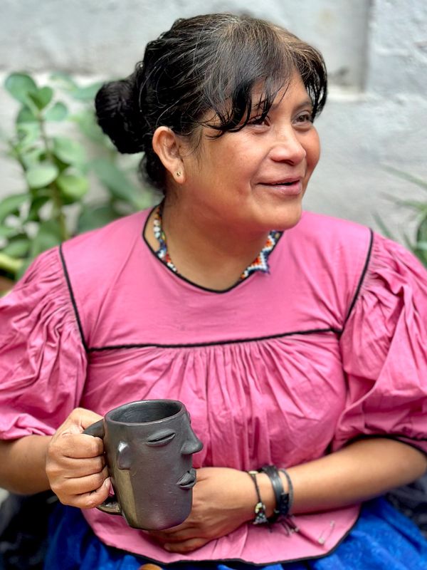 Woman in pink blouse holding a unique face-shaped mug, smiling gently.