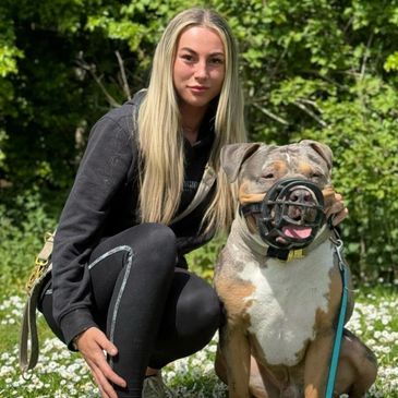 A woman with long blonde hair squats next to a muzzled dog outdoors.