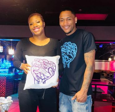 Two people smiling, one holding a pillow with a heart and hands design.