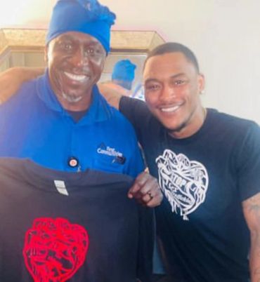 Two smiling men holding a black shirt with a red design indoors.