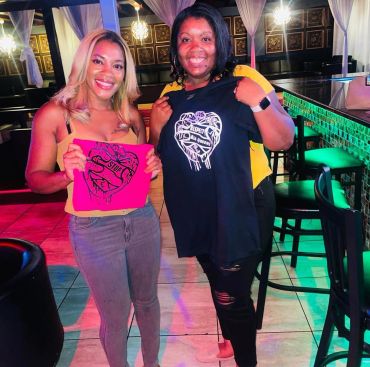 Two women smiling and holding matching shirts with heart designs in a colorful indoor setting.
