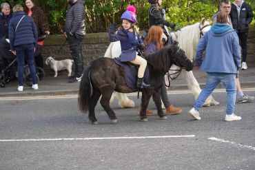 Child riding a small black pony during a public event on a street.