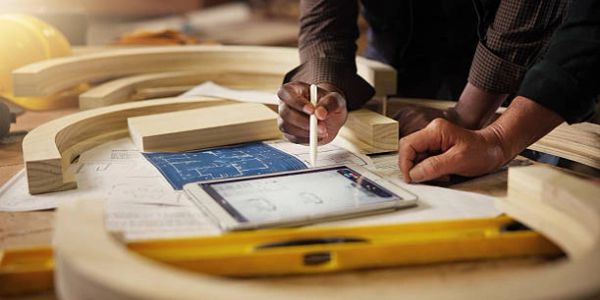 Hands working on architectural plans with tablet and wooden pieces.