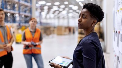 A woman leads a discussion in a warehouse while holding a tablet.