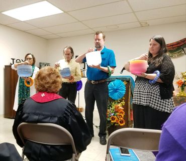 Four people hold colorful bowls in a room with a seated audience.