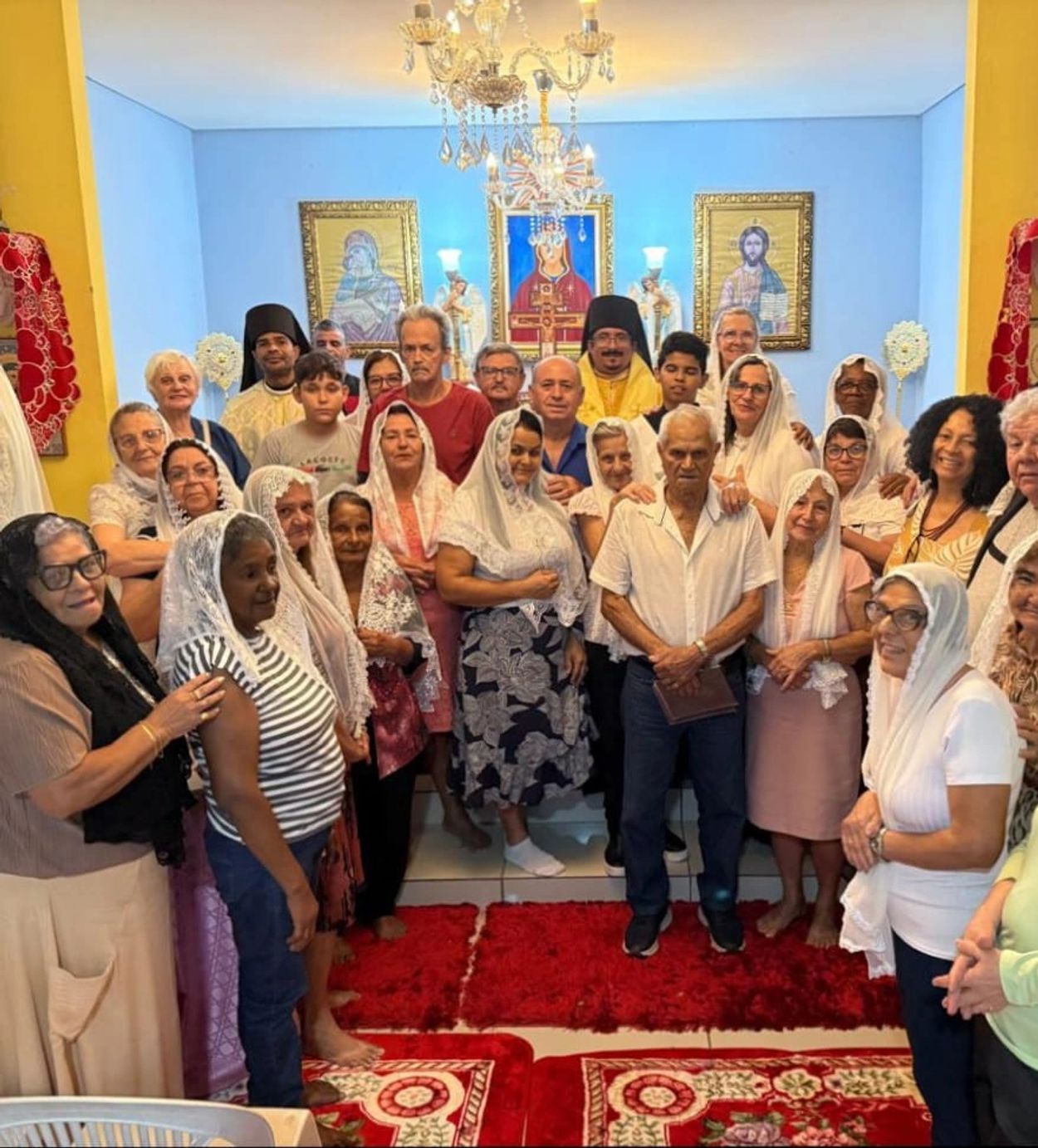 A large group of people gathered inside a room with religious icons and a chandelier.