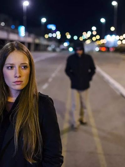 A woman looks concerned as a shadowy figure follows her at night on an empty street.