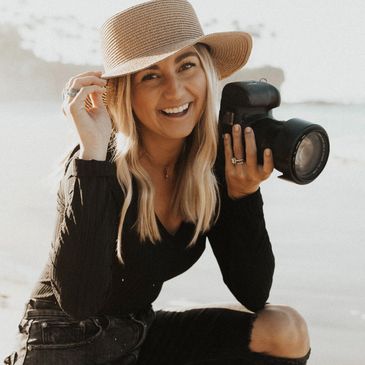 kendra sutton is holding her hat and smiling with her canon camera at the beach