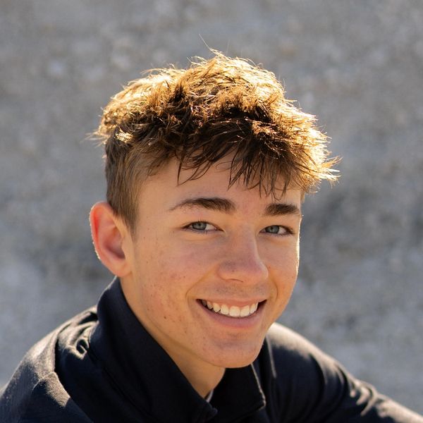 A young man with tousled hair smiling outdoors against a blurred background.