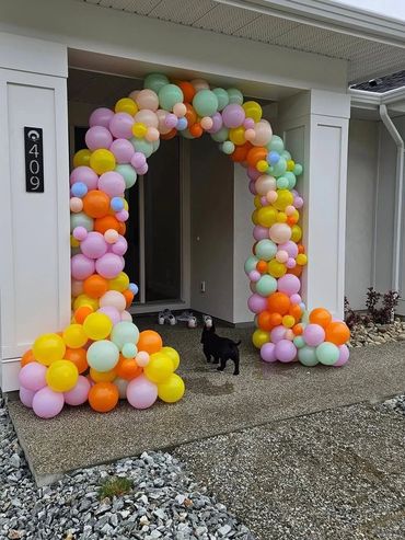 Colorful balloon arch decorates a house entrance with a small black dog nearby.