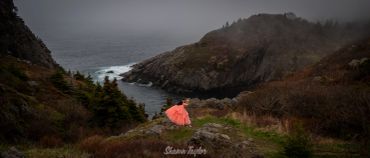 A person in a bright dress stands on a cliff overlooking misty mountains and water.