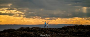 Couple holding hands on rocky shore at sunset.