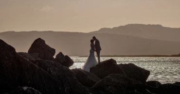 Couple embraces on rocks by the sea at sunset.