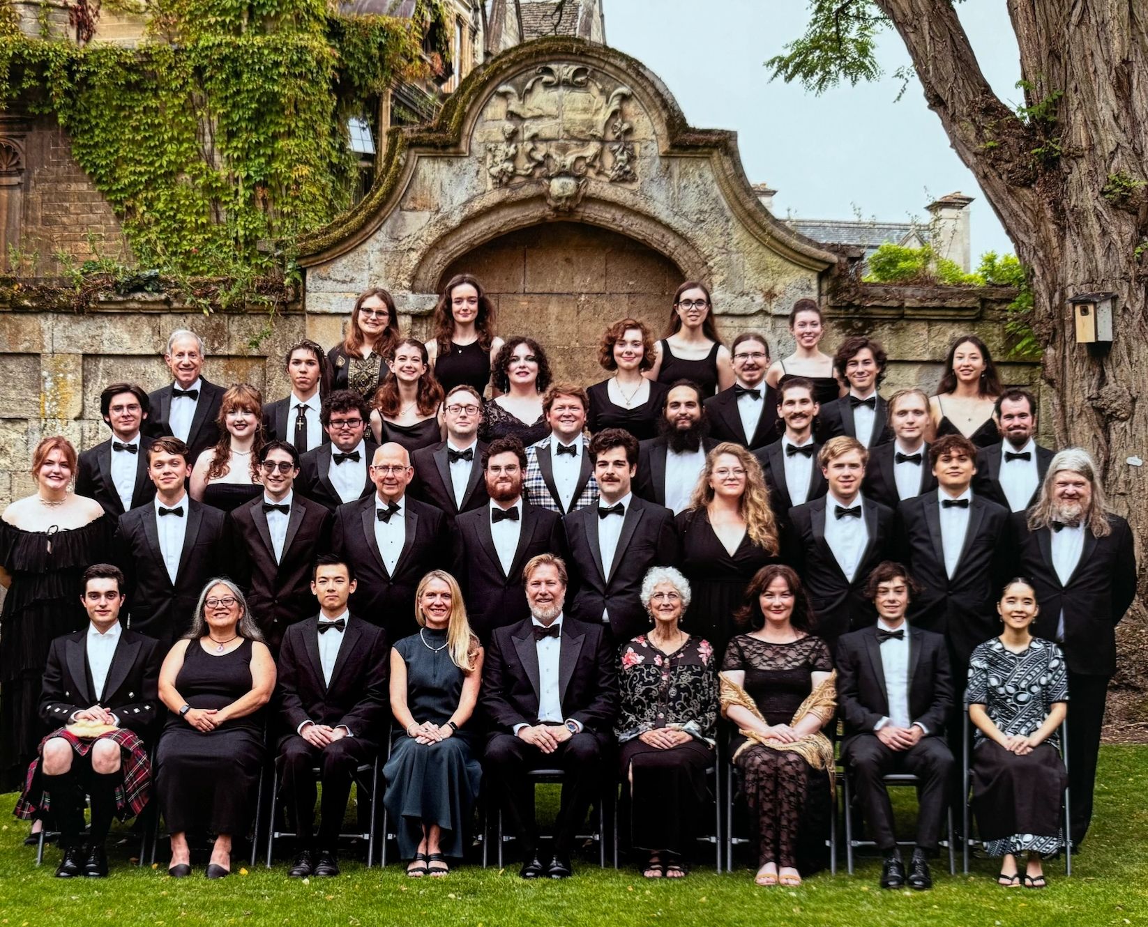 Large formal group photo in tuxedos and dresses outdoors.