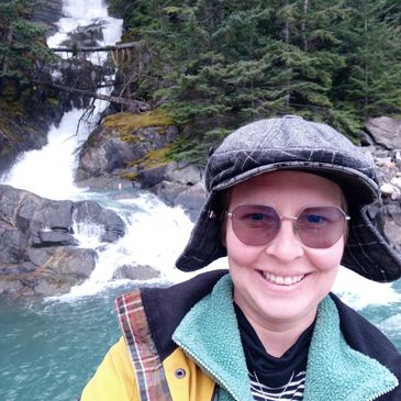 Smiling person in outdoor gear near a waterfall and forest.