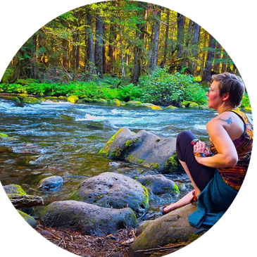 Person sitting contemplatively by a forest river with sunlight filtering through trees.