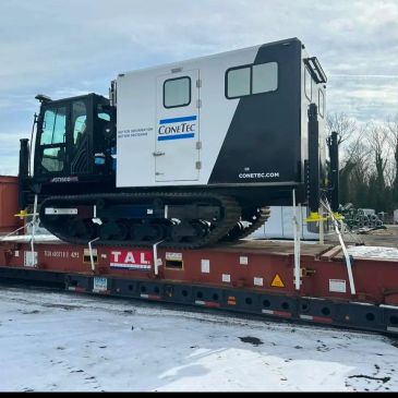 A tracked CONETEC machine loaded on a flatbed trailer in a snowy area.