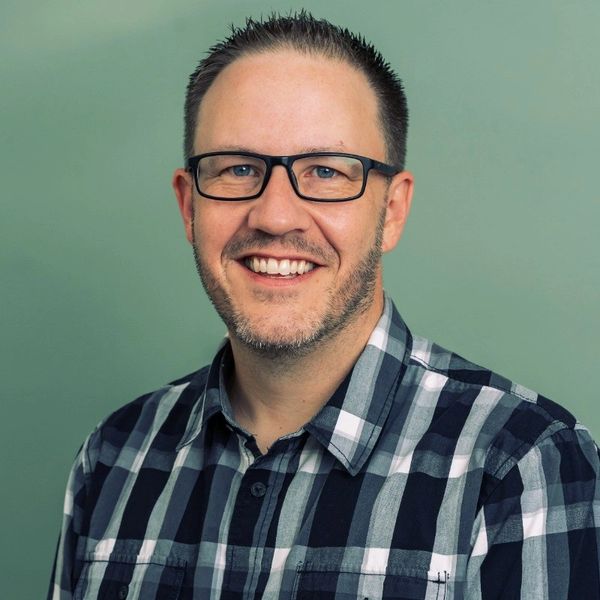 Smiling man wearing glasses and a checkered shirt against a plain background.