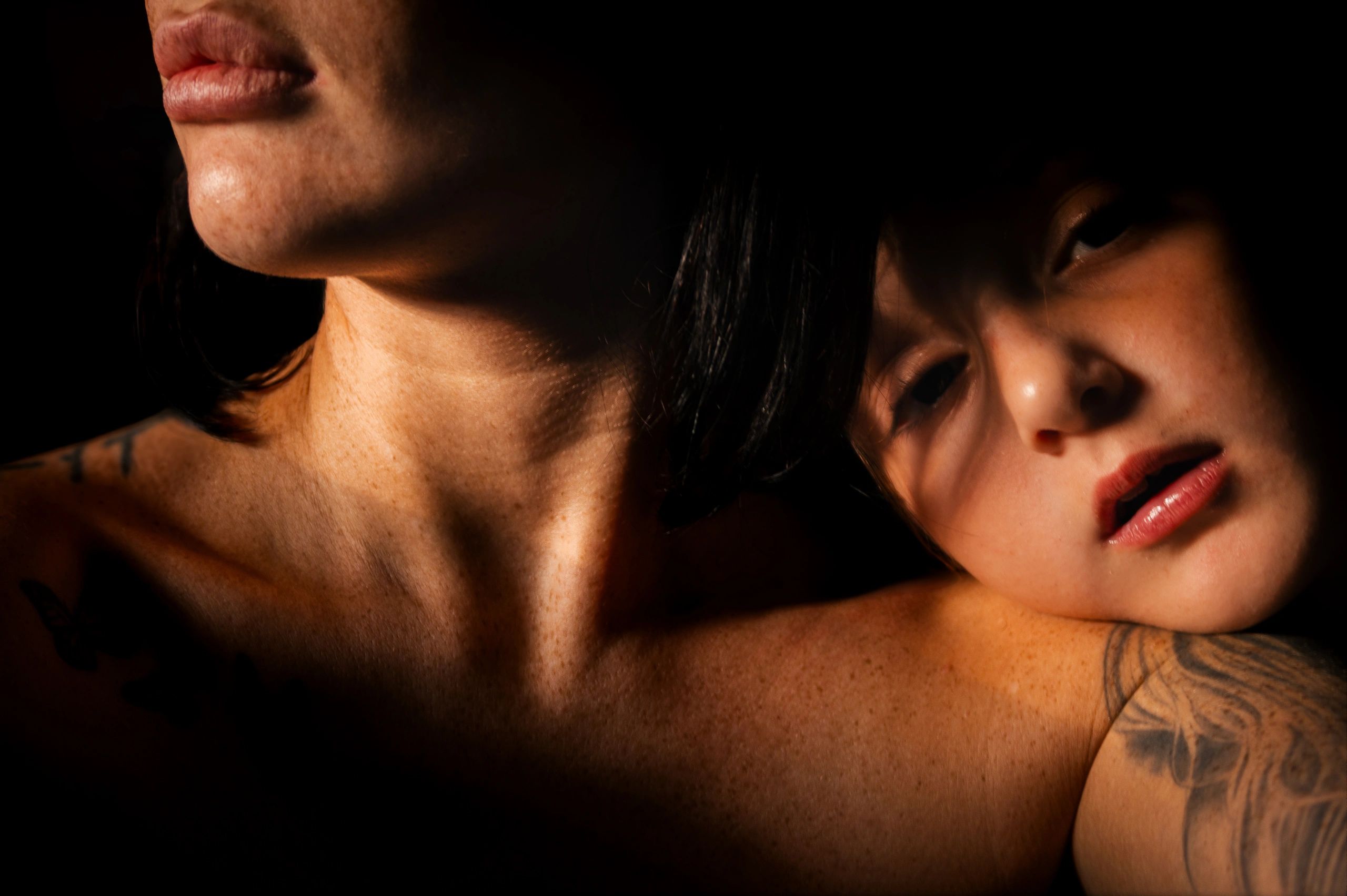 Close-up of two individuals in dramatic lighting, showing skin texture and tattoos.