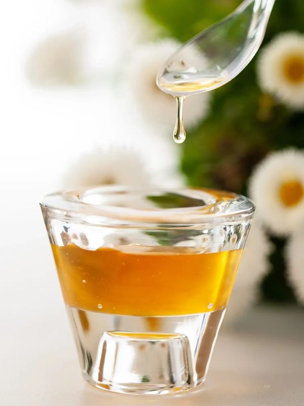 Honey dripping from a spoon into a glass container with flowers in the background.