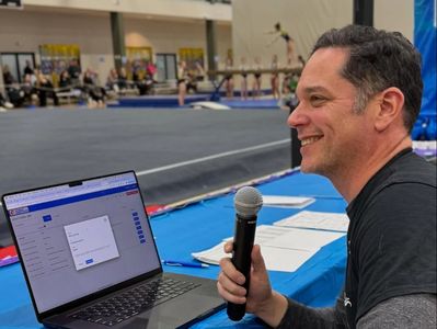 Man holding microphone, judging a gymnastics event with laptop open.