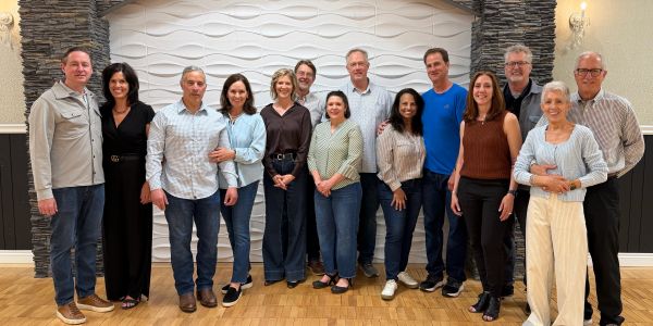 Group of 14 people posing indoors against a textured white wall.