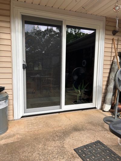 Sliding glass door with screen on a patio, reflecting trees and outdoor furniture.