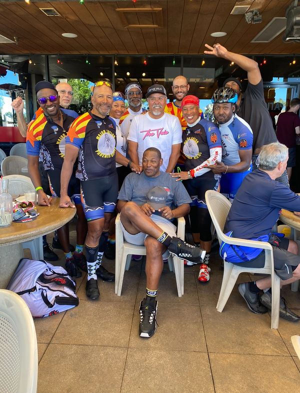 A joyful group of cyclists posing together at a cafe, some in matching Major Taylor jerseys.