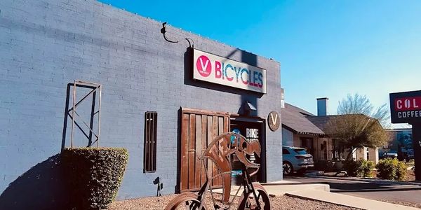 Metal bicycle sculpture outside a V Bicycles shop under clear blue sky.
