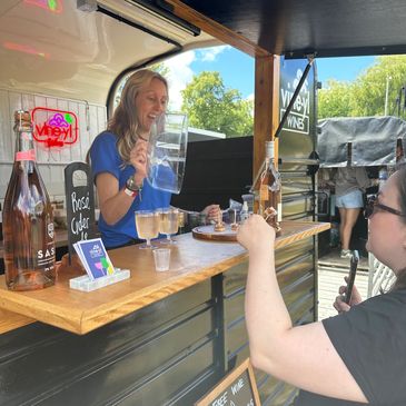 A woman serving wine and tapas samples at an outdoor stand on a sunny day.