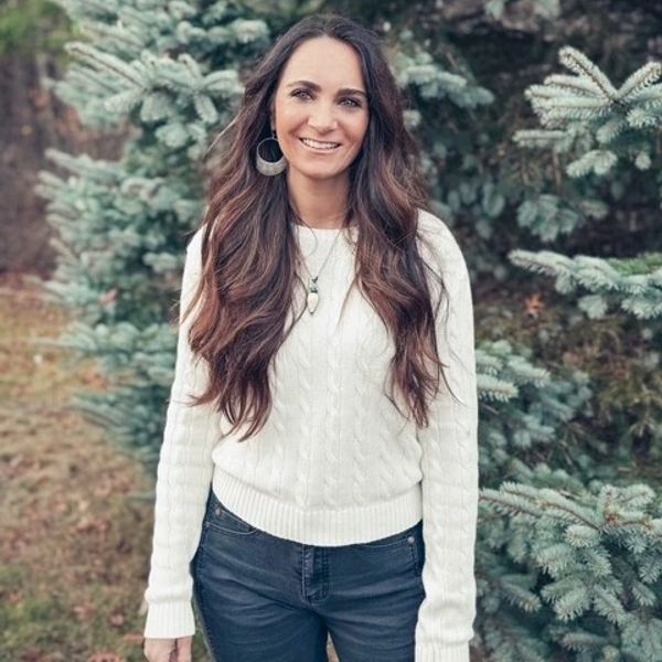 Smiling woman in white sweater stands outdoors near evergreen trees.