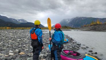 Two kayakers stand by a river with mountains in the background.
