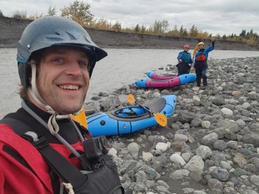 Three people with kayaks on a rocky riverbank, smiling and celebrating.