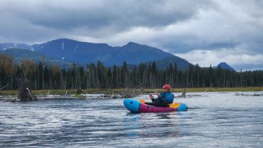 Person kayaking on a calm lake with mountains and cloudy sky in the background.