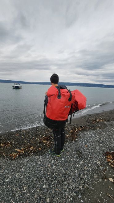 Person in wetsuit with large red bag standing on rocky beach looking at boat.
