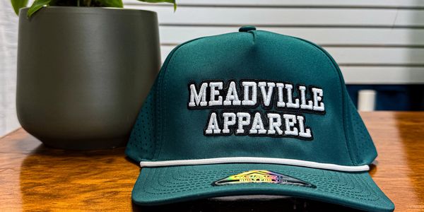 Green Meadville Apparel cap on wooden table next to a plant.