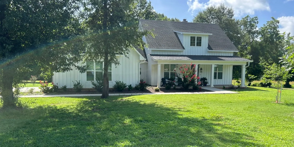 A white house with a porch surrounded by lush green lawn and trees under a clear blue sky.