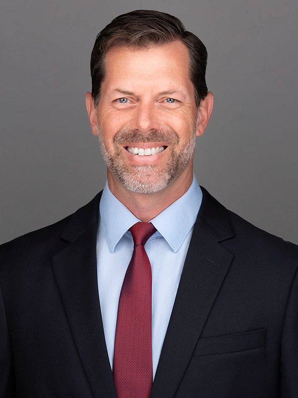Smiling middle-aged man in a suit and red tie against a gray background.