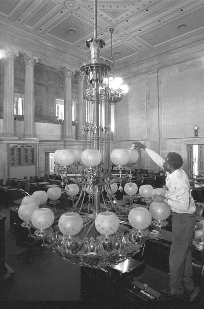 Man cleaning a large chandelier inside a grand, classical-style room.