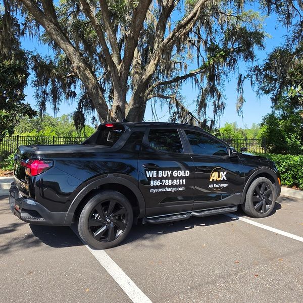 Black pickup truck with gold buying service advertisement parked under a large tree.